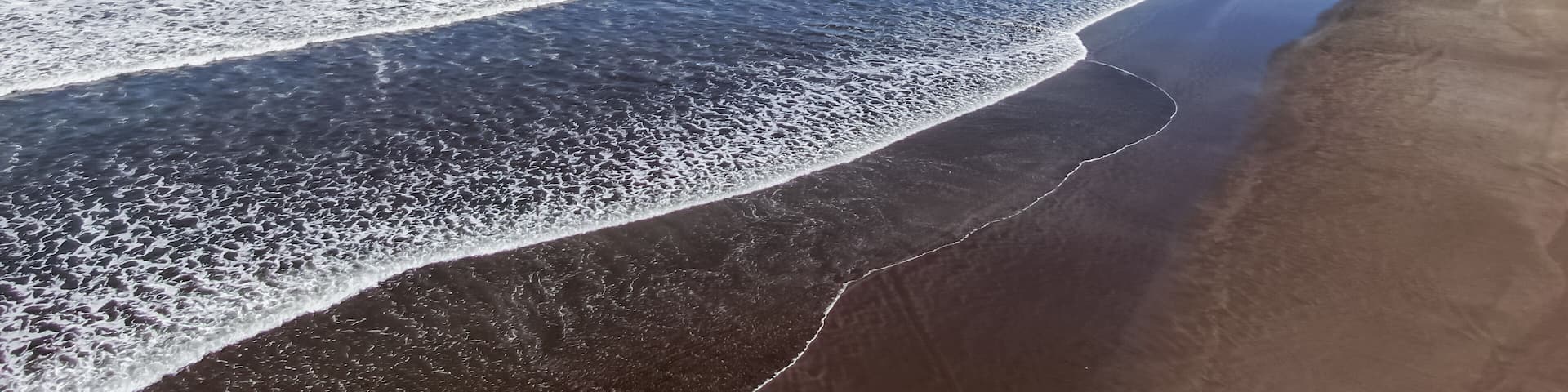 A beach with waves, a rock formation, and mountains in the background.
