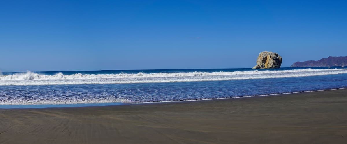 A beach with waves, a large rock formation, and distant mountains.