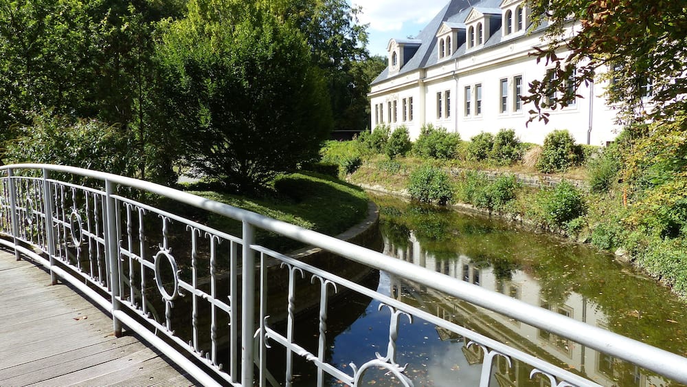 Un ponte con ringhiere in metallo elaborate, un fiume tranquillo, vegetazione rigogliosa e un edificio classico con un tetto ripido.