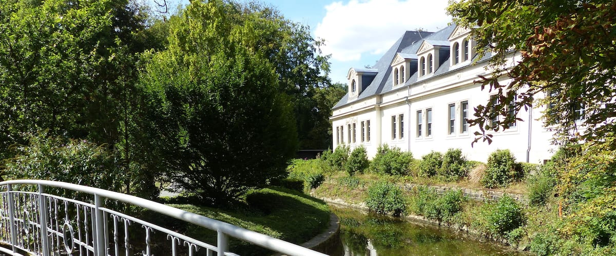 A bridge with ornate metal railings, a calm river, lush greenery, and a classical building with a steep roof.