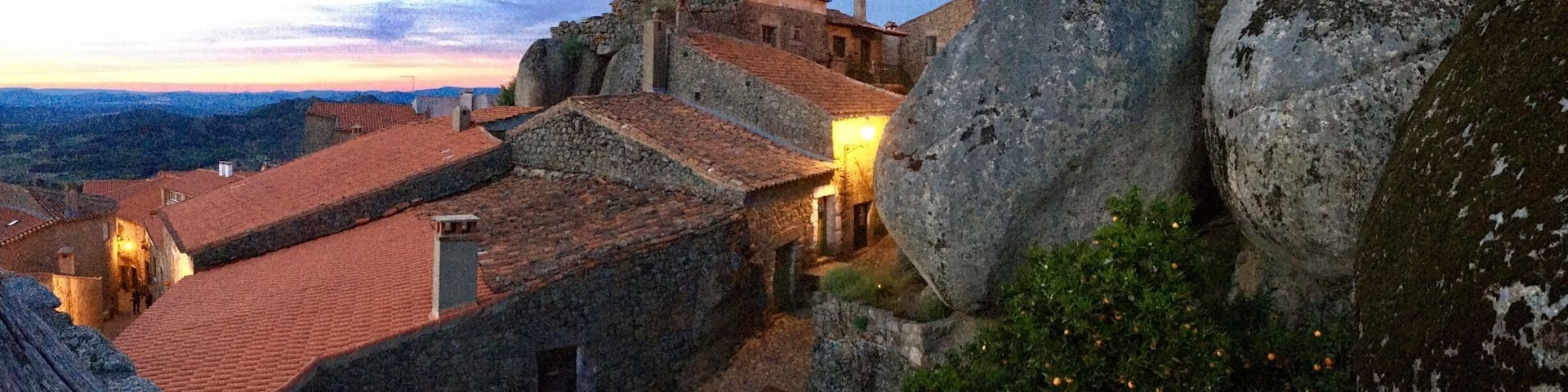 A historic stone building with red-tiled roofs, large boulders, and a sunset in the background.