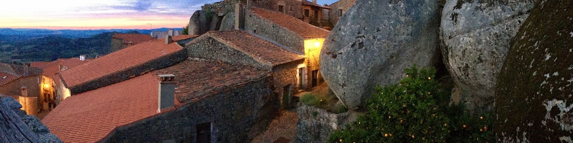 Um edifício histórico de pedra com telhados de telhas vermelhas, grandes pedras e um pôr do sol ao fundo.