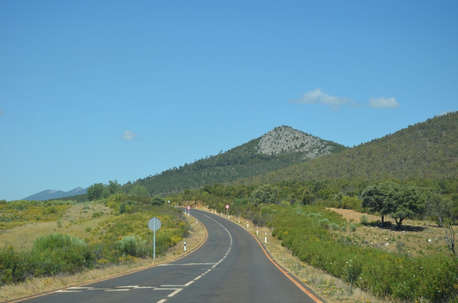 Uma estrada sinuosa serpenteia por uma paisagem montanhosa com vegetação escassa e uma montanha ao fundo.