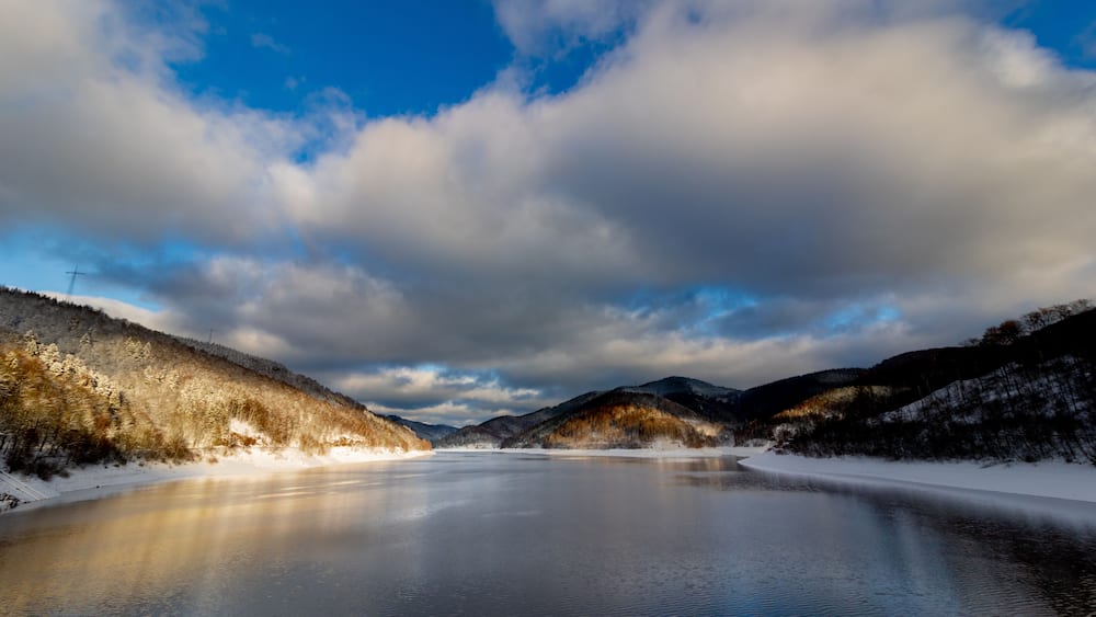 A frozen lake surrounded by snow-covered hills and a partly cloudy sky.