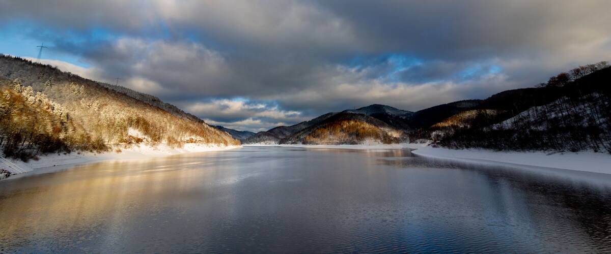 A frozen lake surrounded by snow-covered hills and a partly cloudy sky.