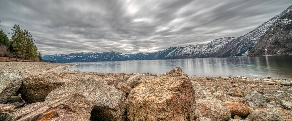 A rocky shoreline with a calm lake, snow-capped mountains in the background, and a cloudy sky.