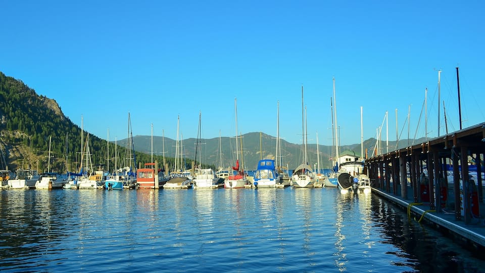 A marina with various sailboats docked along a wooden pier, surrounded by a mountainous landscape.