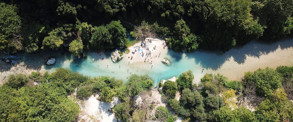 An aerial view of a beach surrounded by dense forest, with clear turquoise waters and rocky formations.