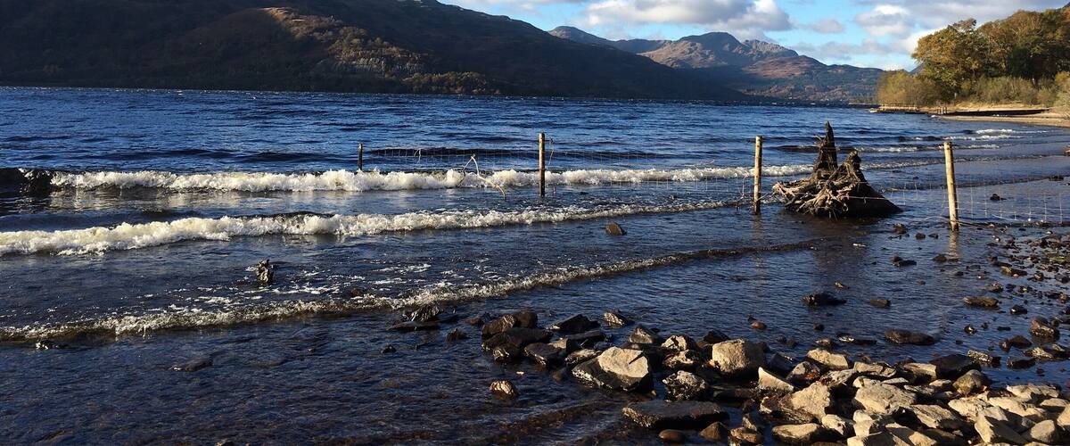 A rocky shoreline with waves, a wooden fence, and mountains in the background.