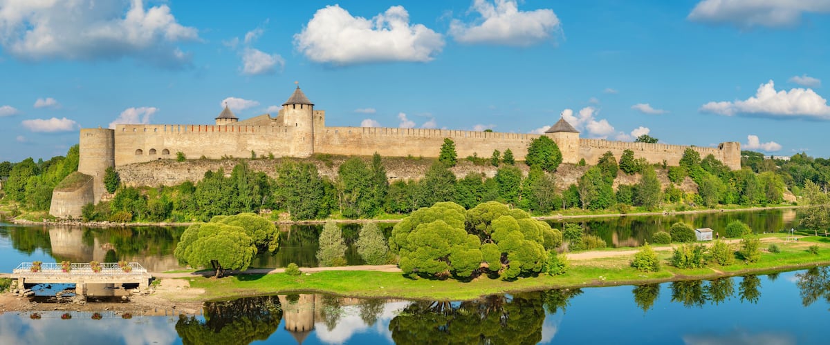 A historic castle by a calm river with lush greenery and a clear sky.