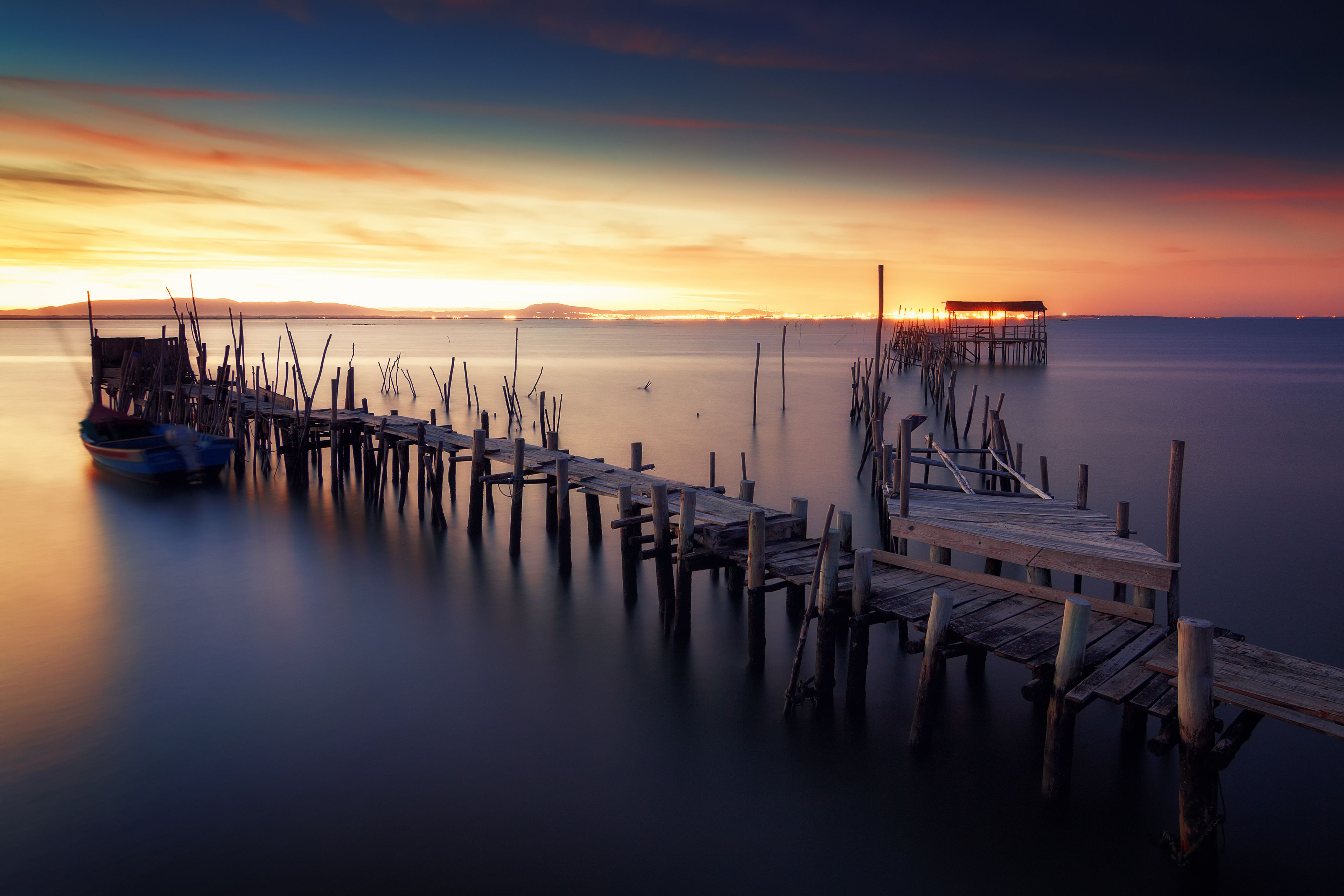 A wooden pier extending into calm waters at sunset with a boat docked on the left and a small hut on the right.