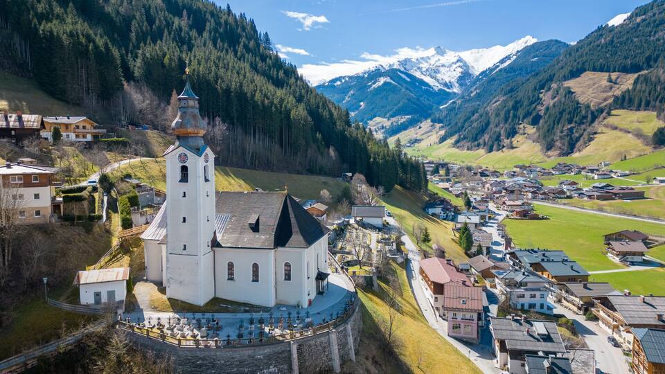A white church with a bell tower, surrounded by a village and mountains in the background.
