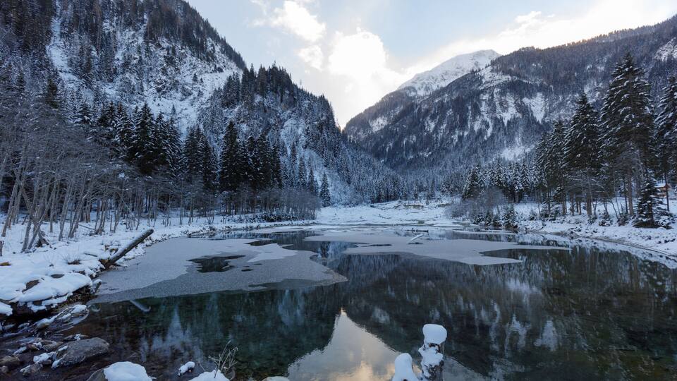 A snow-covered mountain landscape with a clear lake reflecting the surrounding trees and mountains.