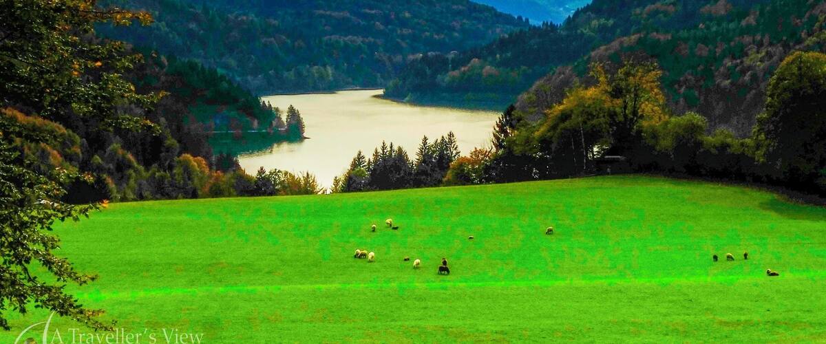 A valley with a lake, green fields, and snow-capped mountains in the background.