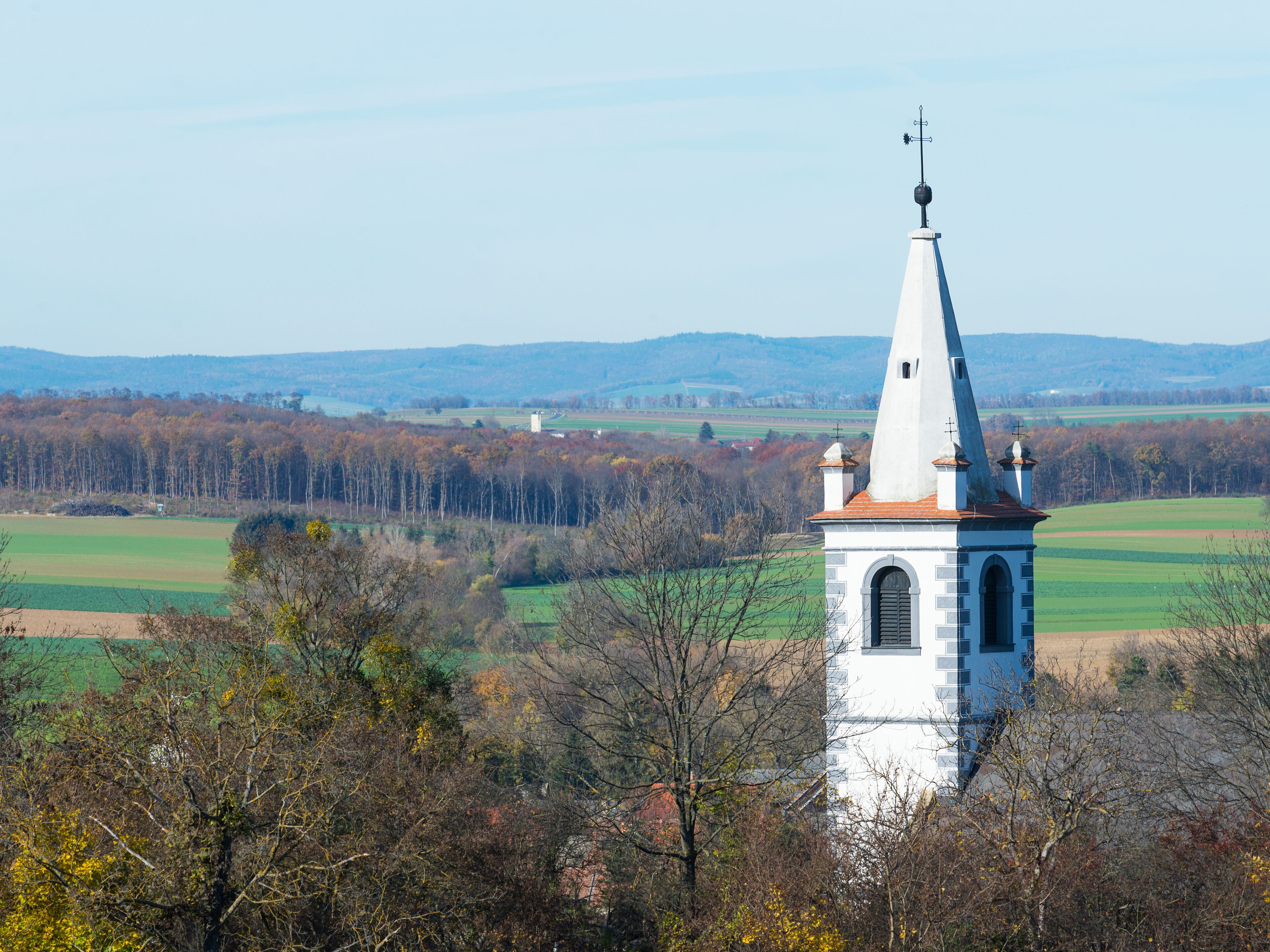 A church with a tall steeple and a weather vane, surrounded by autumn-colored trees and fields.