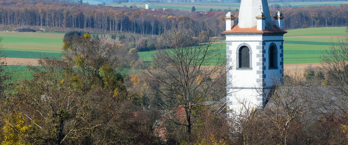 A church with a tall steeple and a weather vane, surrounded by autumn-colored trees and fields.