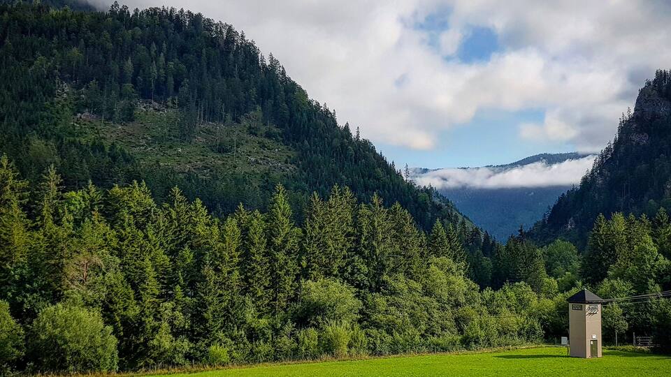 A lush green field with a small building in the foreground, surrounded by dense forest and mountains under a partly cloudy sky.