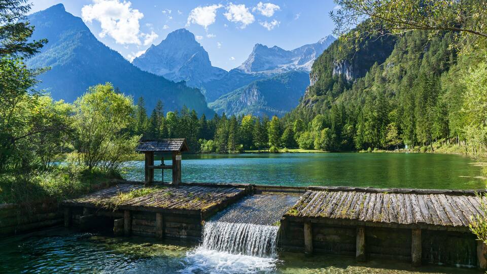 A wooden structure with a small waterfall, surrounded by lush green mountains and a calm lake.