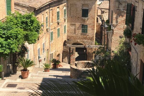 Una stretta via fiancheggiata da edifici con tetti in terracotta, piante in vaso e vista sulle dolci colline in lontananza.