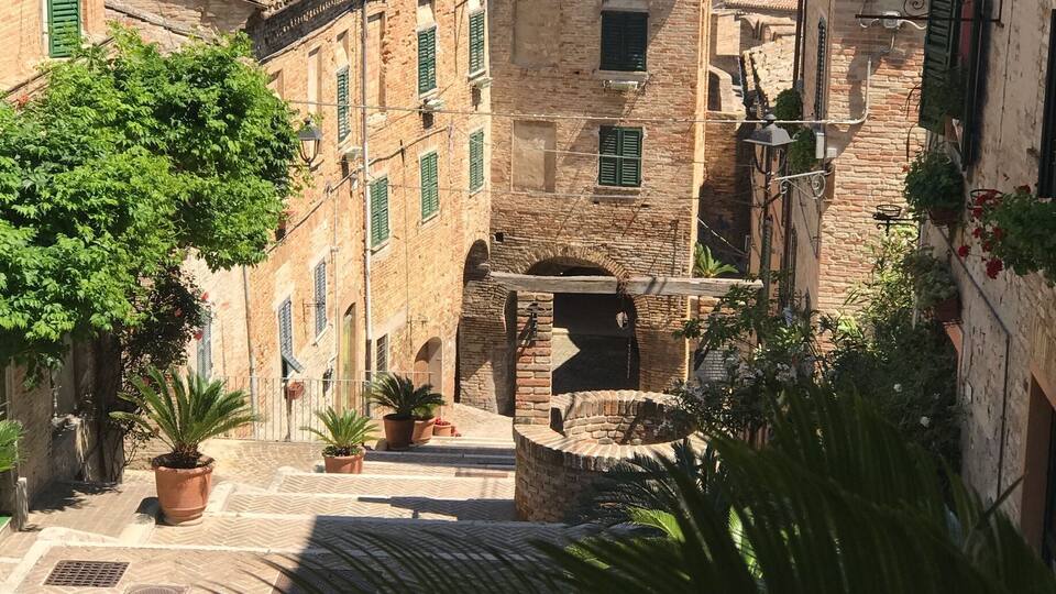 A narrow street with terracotta-tiled buildings, potted plants, and a view of rolling hills in the distance.