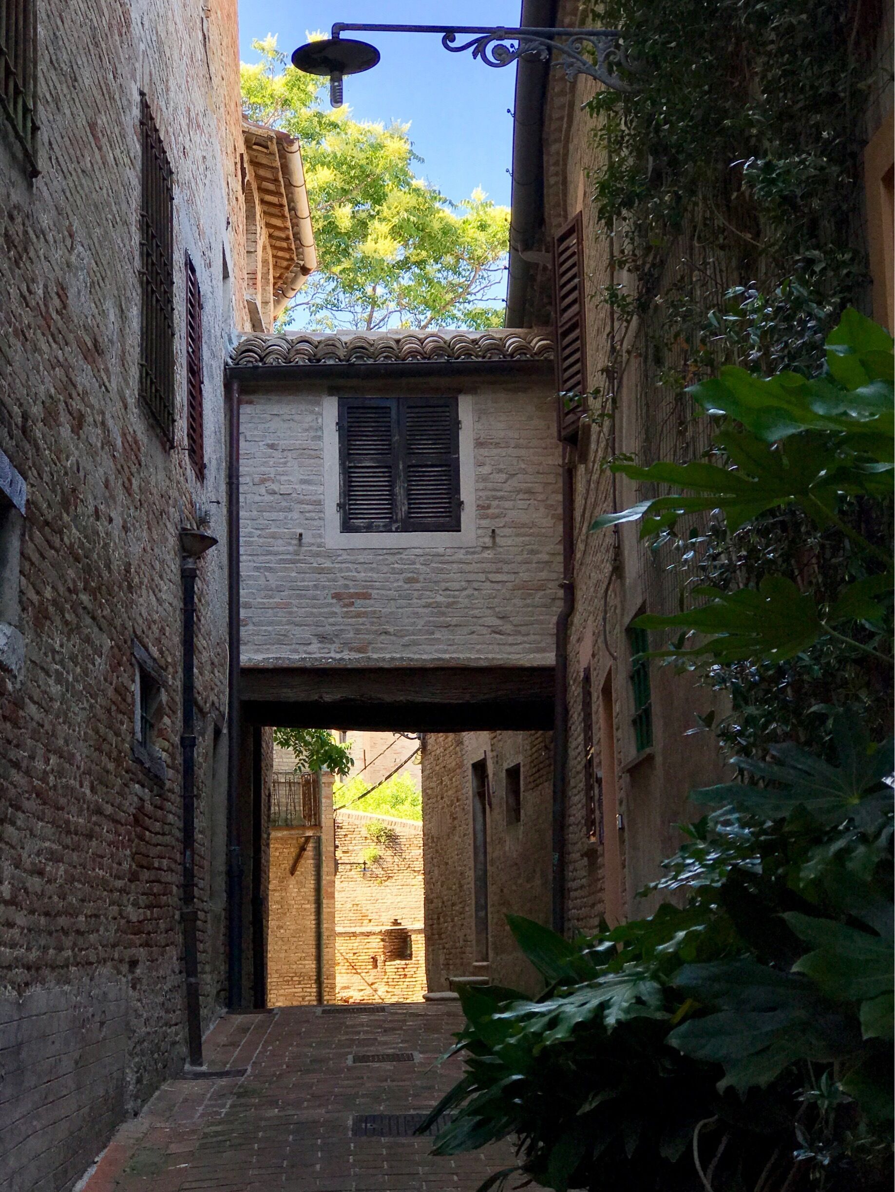 A narrow alleyway with brick buildings, a small window with shutters, and a street lamp.
