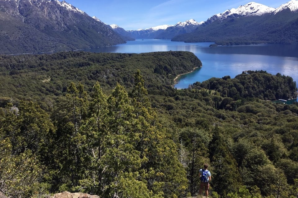 A hiker on a mountain trail with a lake and snow-capped mountains in the background.
