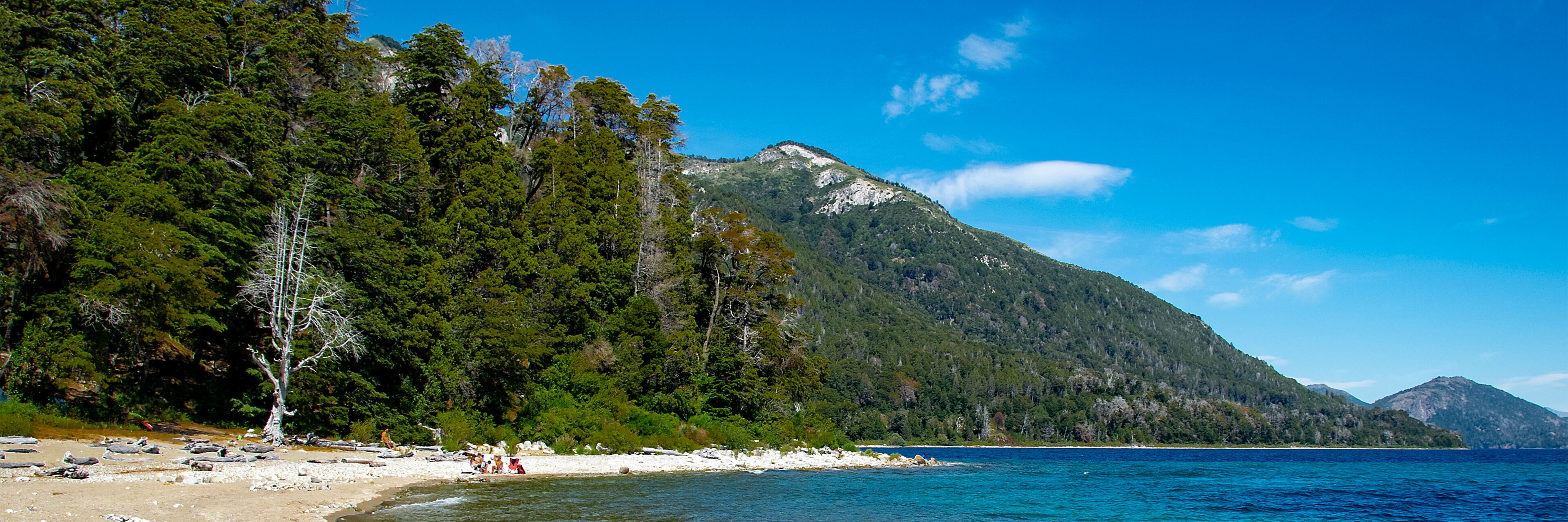 Uma praia com areia branca, costa rochosa, floresta verde densa e uma montanha ao fundo.