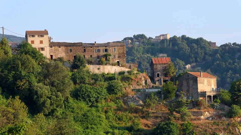 A hillside village with historic stone buildings, lush greenery, and a prominent hilltop structure.