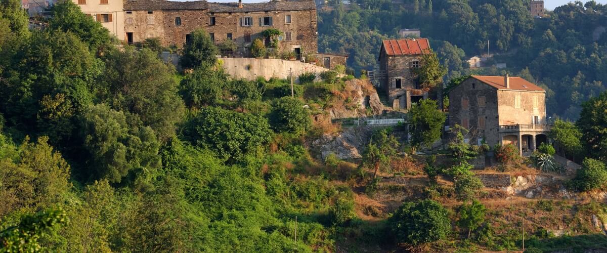A hillside village with historic stone buildings, lush greenery, and a prominent hilltop structure.