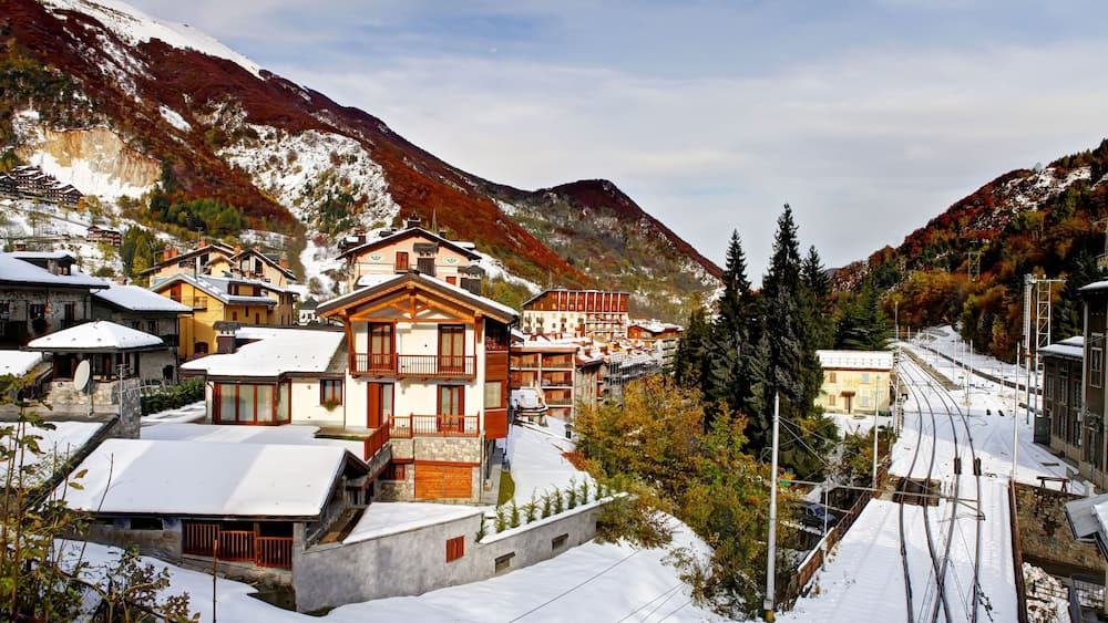 Un village enneigé, avec ses maisons traditionnelles en bois, une voie ferrée et des montagnes en arrière-plan.