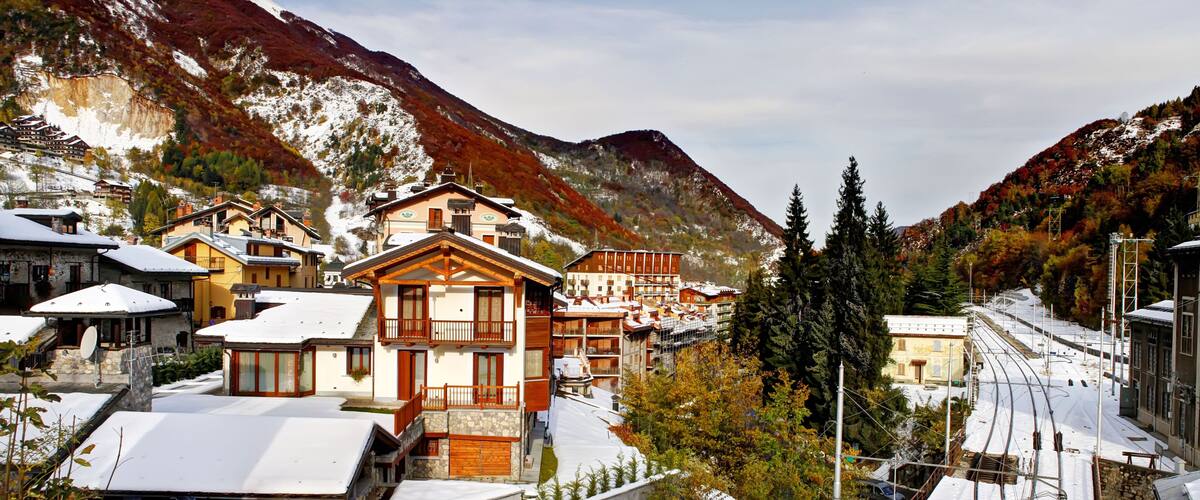 A snow-covered village with traditional wooden houses, a railway track, and mountains in the background.