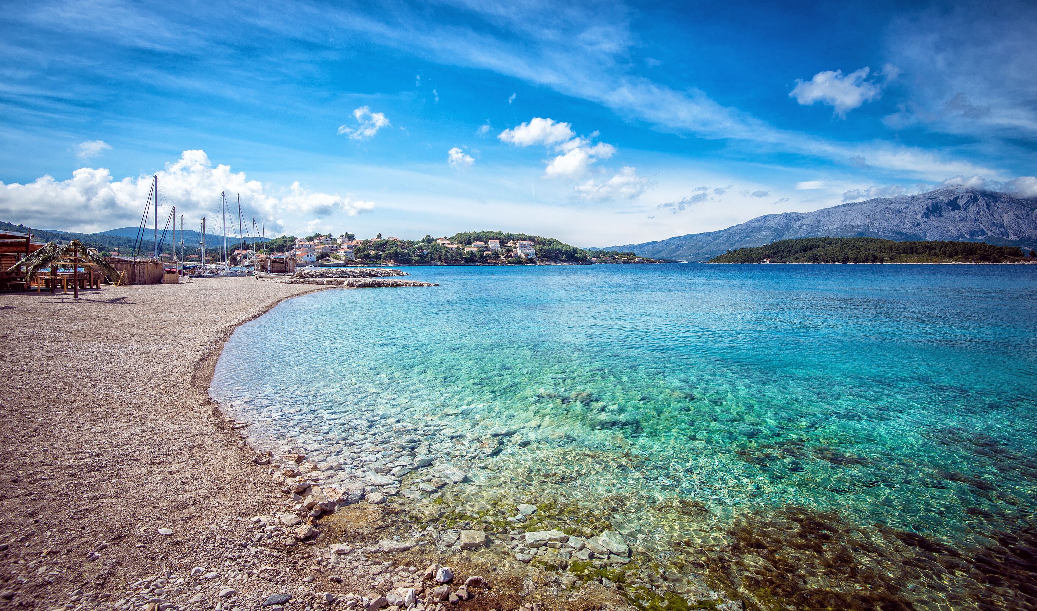 A clear coastal beach with rocky shore, a small settlement, and mountains in the background.