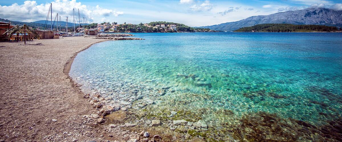 A clear coastal beach with rocky shore, a small settlement, and mountains in the background.