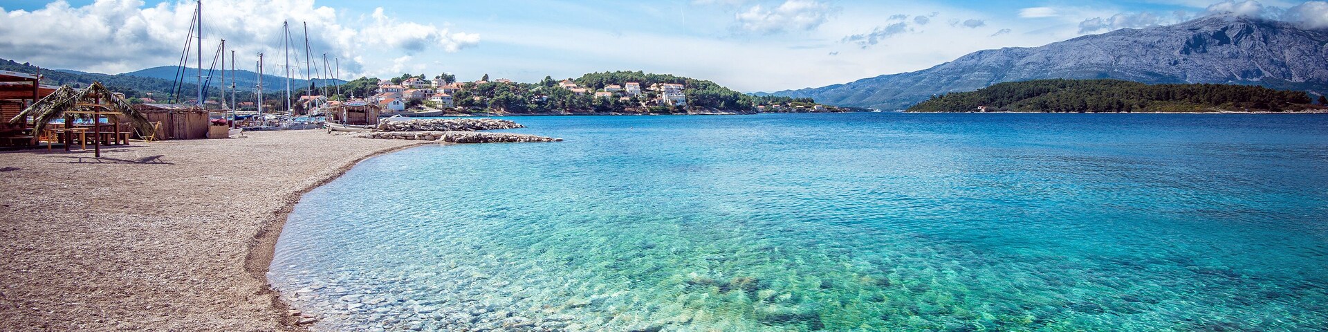 A clear coastal beach with rocky shore, a small settlement, and mountains in the background.