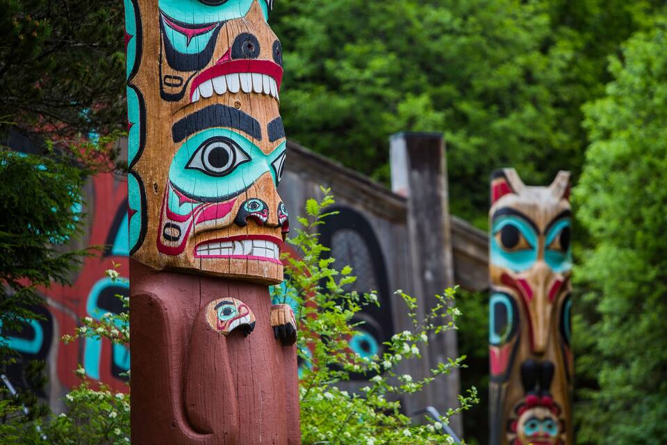Two totem poles with intricate carvings and a lush green forest background.