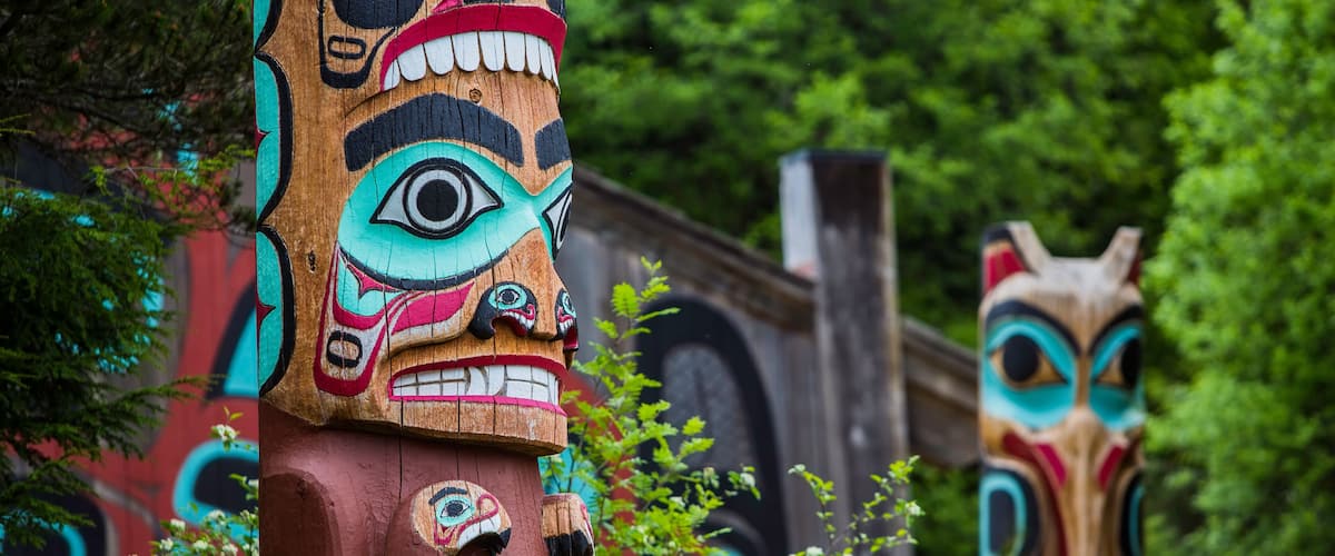 Two totem poles with intricate carvings and a lush green forest background.