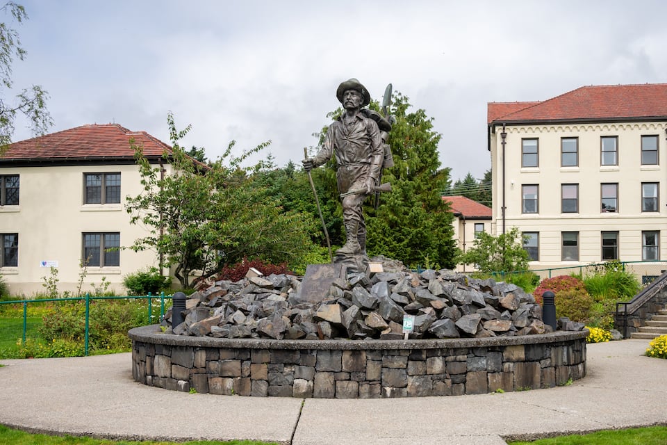 A statue of a man with a rifle on a stone pedestal in a park with buildings in the background.