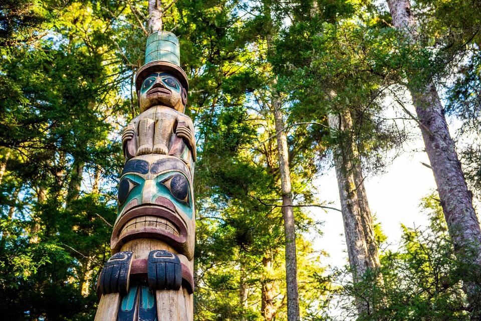 A totem pole with intricate carvings, set against a backdrop of tall trees and sunlight filtering through the foliage.