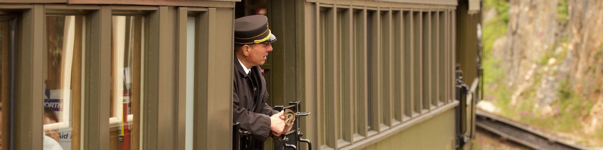 Un tren con las letras 'R' y 'W' en la carrocería, un hombre con uniforme sosteniendo un billete y un paisaje montañoso al fondo.