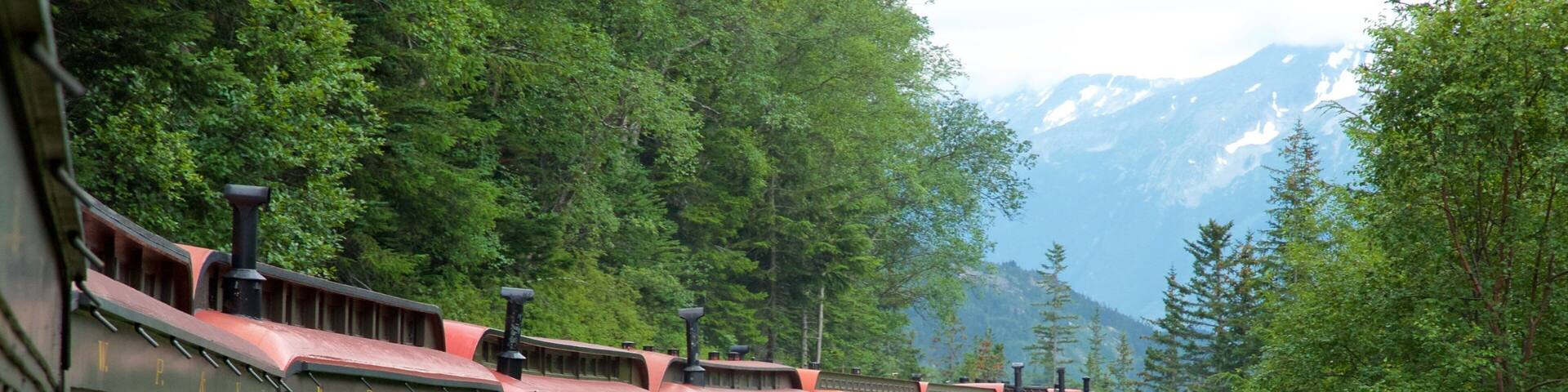 A vintage train with 'Lake Fraser' written on the side, traveling through a forested area with mountains in the background.