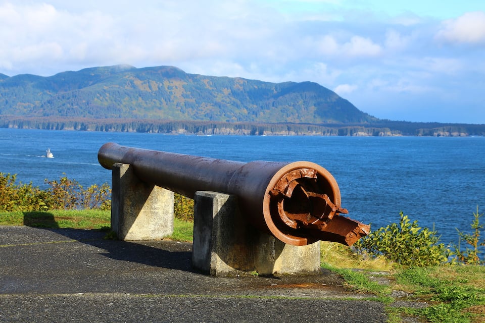 A cannon on a concrete platform by a lake with mountains in the background.