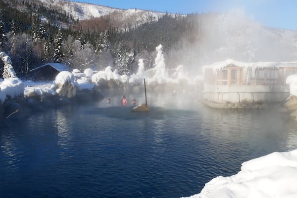 A hot spring with steam rising, surrounded by snow-covered trees and a wooden cabin.