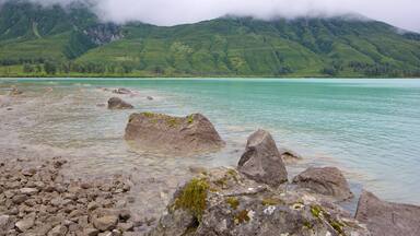 A lake with clear turquoise water, surrounded by rocky shores and green hills.