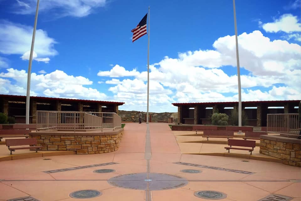 A plaza with a central walkway, benches, and two flags on flagpoles.