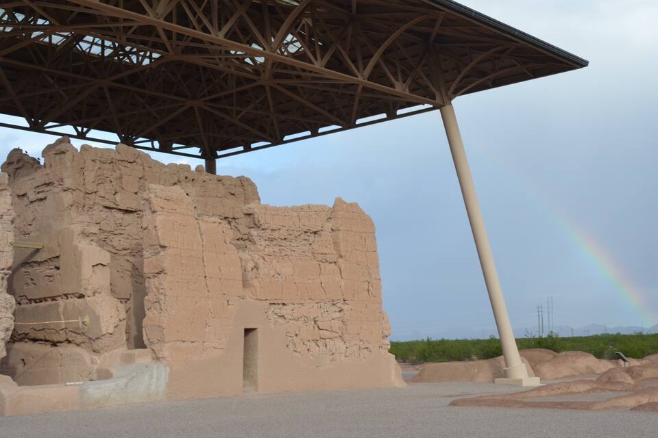 A weathered stone structure with a metal roof, a rainbow in the background, and a clear sky.