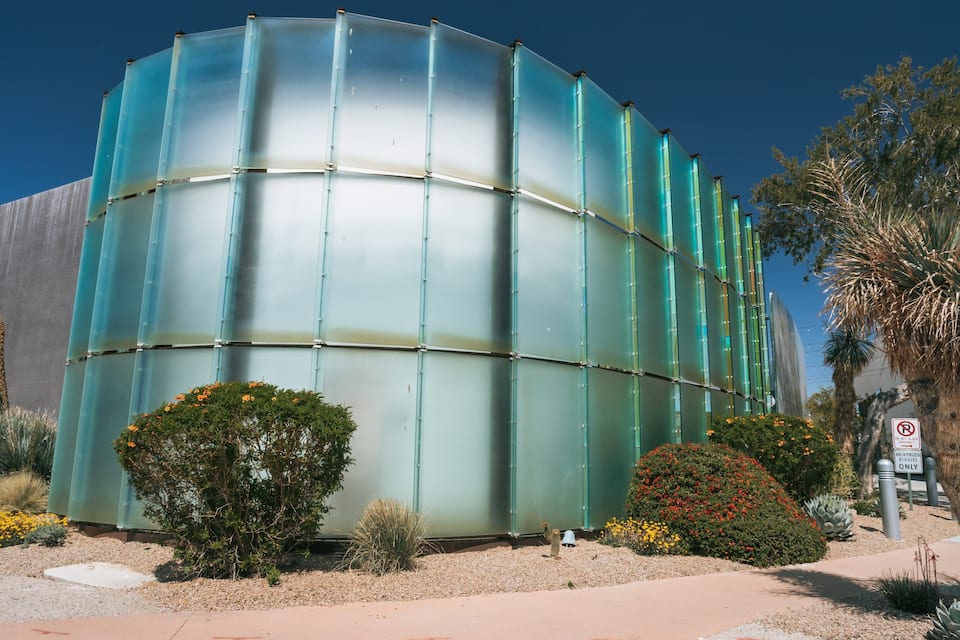 A cylindrical building with a reflective surface, surrounded by desert vegetation and a palm tree.