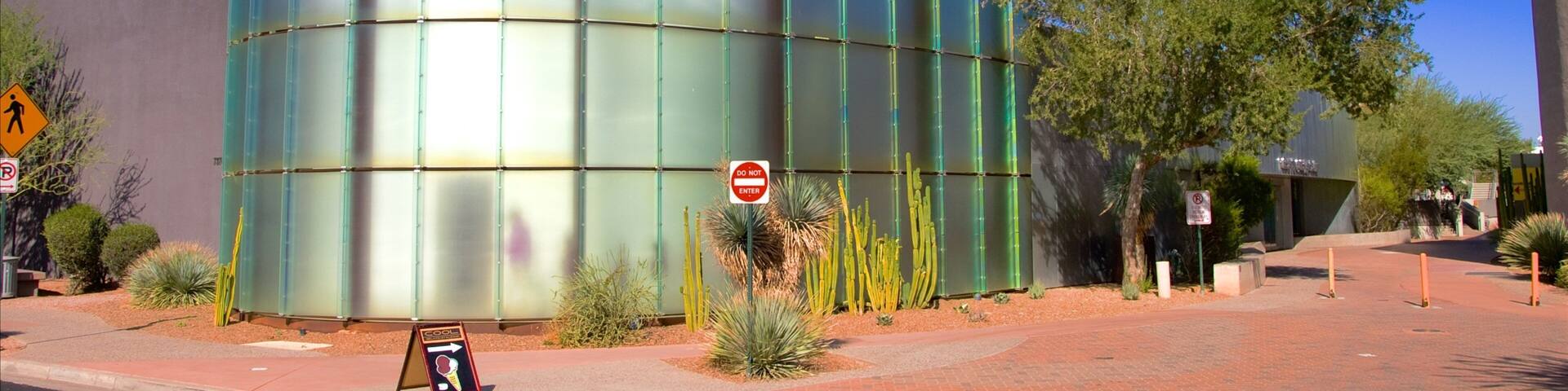 Un edificio moderno con superficie reflectante, rodeado de vegetación desértica y un cielo azul despejado.