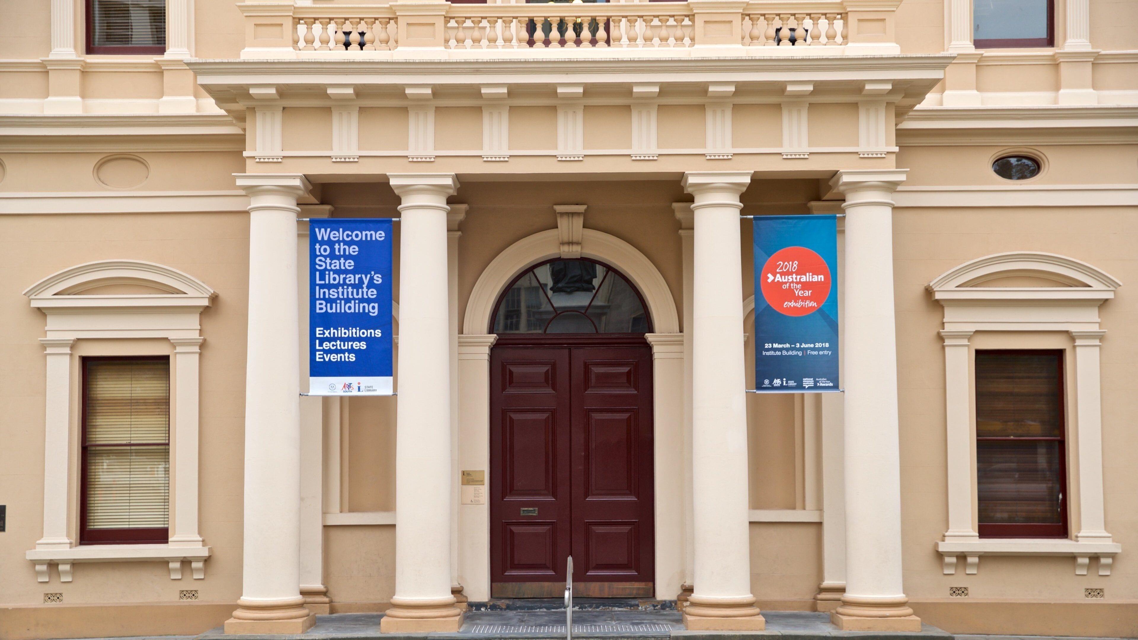 Een gevel met een centrale ingang, geflankeerd door twee kolommen en een balustrade. Twee spandoeken zijn zichtbaar; één heet bezoekers welkom in het Institute Building van de State Library, de ander adverteert de tentoonstelling 'Australian of the Year 2018'.