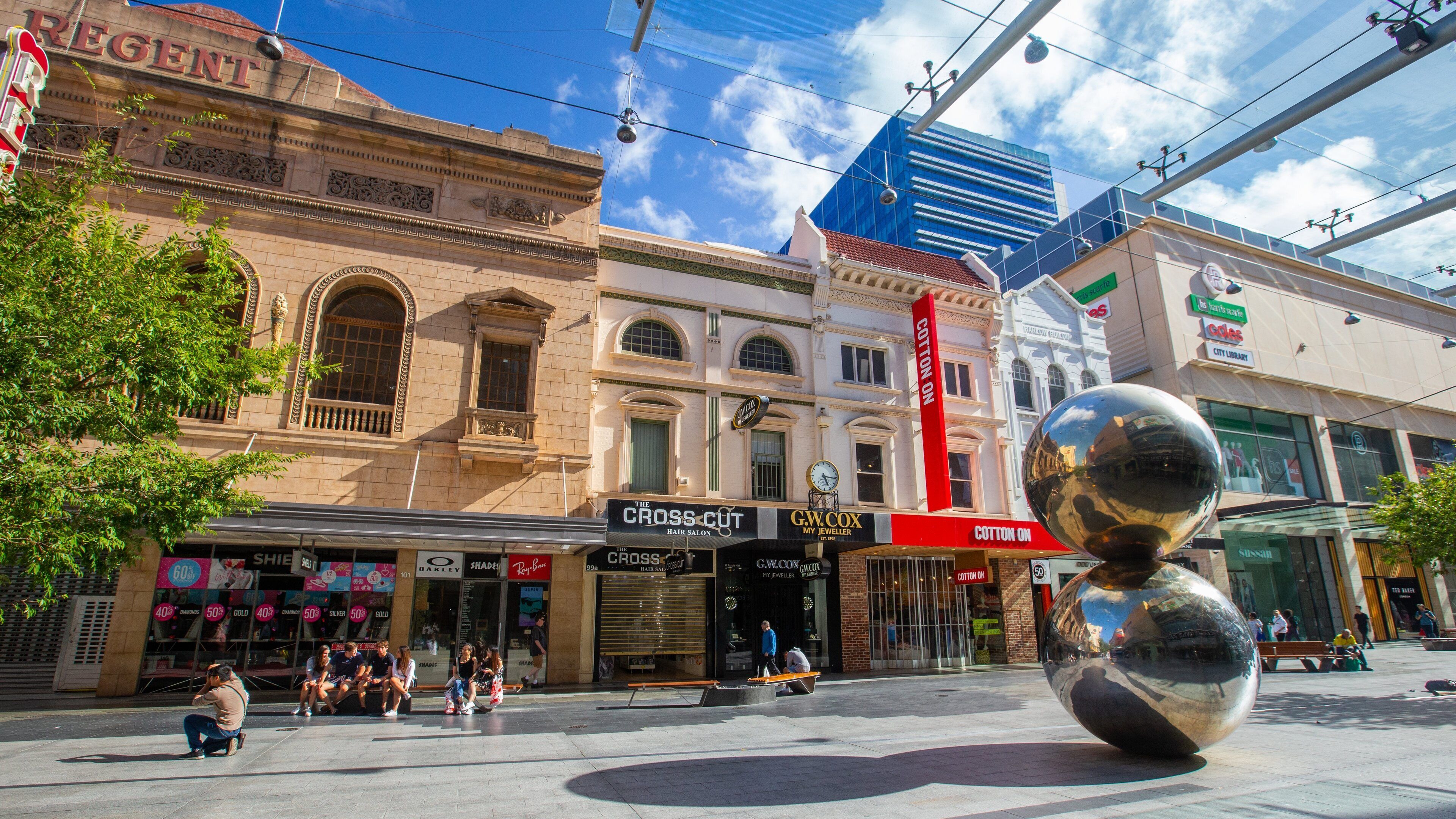 A city square with historic buildings, a large reflective sculpture, and various shops including a hair salon and a jeweler.