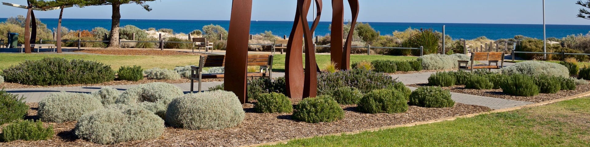 A park with abstract metal sculptures, a large pine tree, and benches along a path with a view of the ocean.